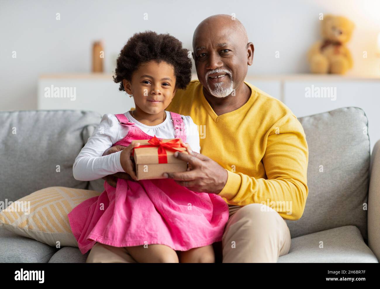 Allegra ragazza afroamericana si siede sulle ginocchia dell'uomo anziano e tiene il regalo dal suo nonno nell'interno del soggiorno. Festeggiamo il compleanno, fami Foto Stock