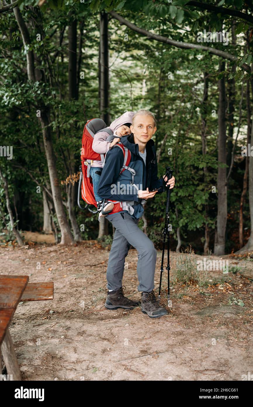 Uomo a piedi con bambino sonnolento in montagna. Bambino seduto in zaino sul retro del padre. Avventura attiva in famiglia nella foresta con ragazza stanca. Freddo Foto Stock