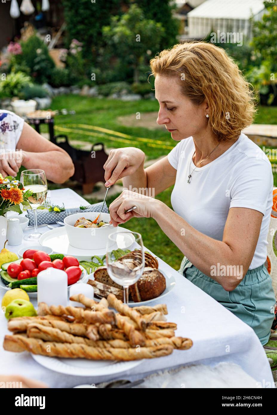 Famiglia felice che ha una cena o un barbecue festivo nel giardino estivo. Giovane bella donna che mangia carne e verdure deliziose al barbecue. Famiglia din Foto Stock