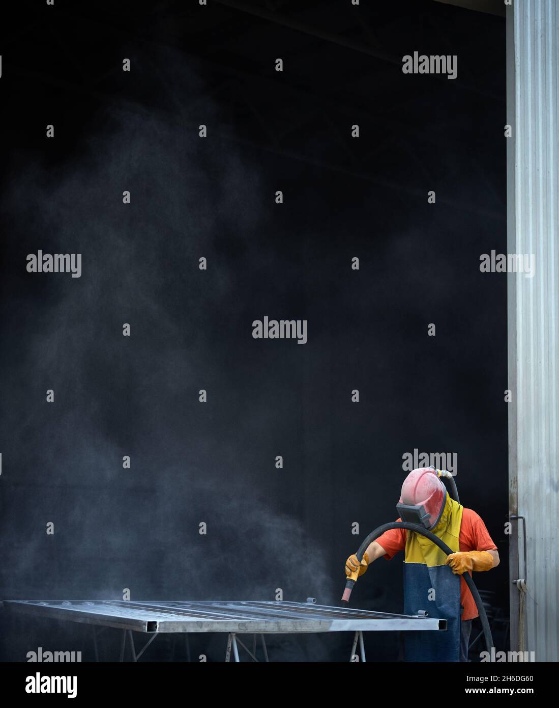 Operatore maschio della sabbiatrice che indossa l'uniforme protettiva durante la lucidatura del metallo in officina. Uomo che prepara i dettagli per la pittura. Foto Stock