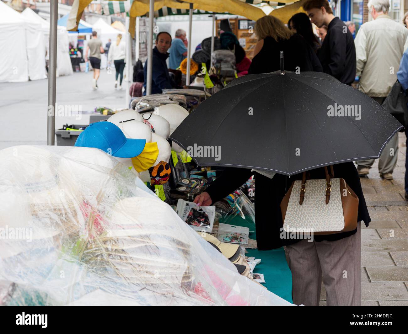 Vecchia donna nascosta sotto un ombrello che guarda i cappelli su una bancarella di mercato, Lymington, Hants, UK Foto Stock