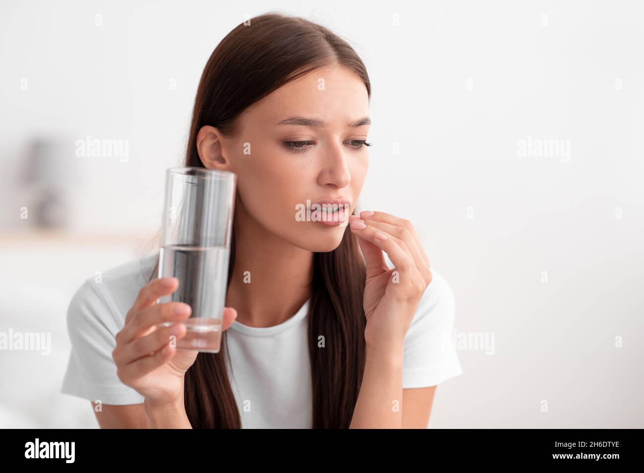 Ritratto di seria giovane donna europea prende la pillola e tiene un bicchiere d'acqua in bianco camera da letto interno Foto Stock