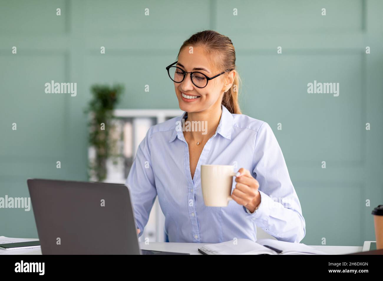 Buona giornata di lavoro. Un lavoratore felice in ufficio che lavora su un laptop e beve caffè, seduto sul posto di lavoro e sorridente Foto Stock
