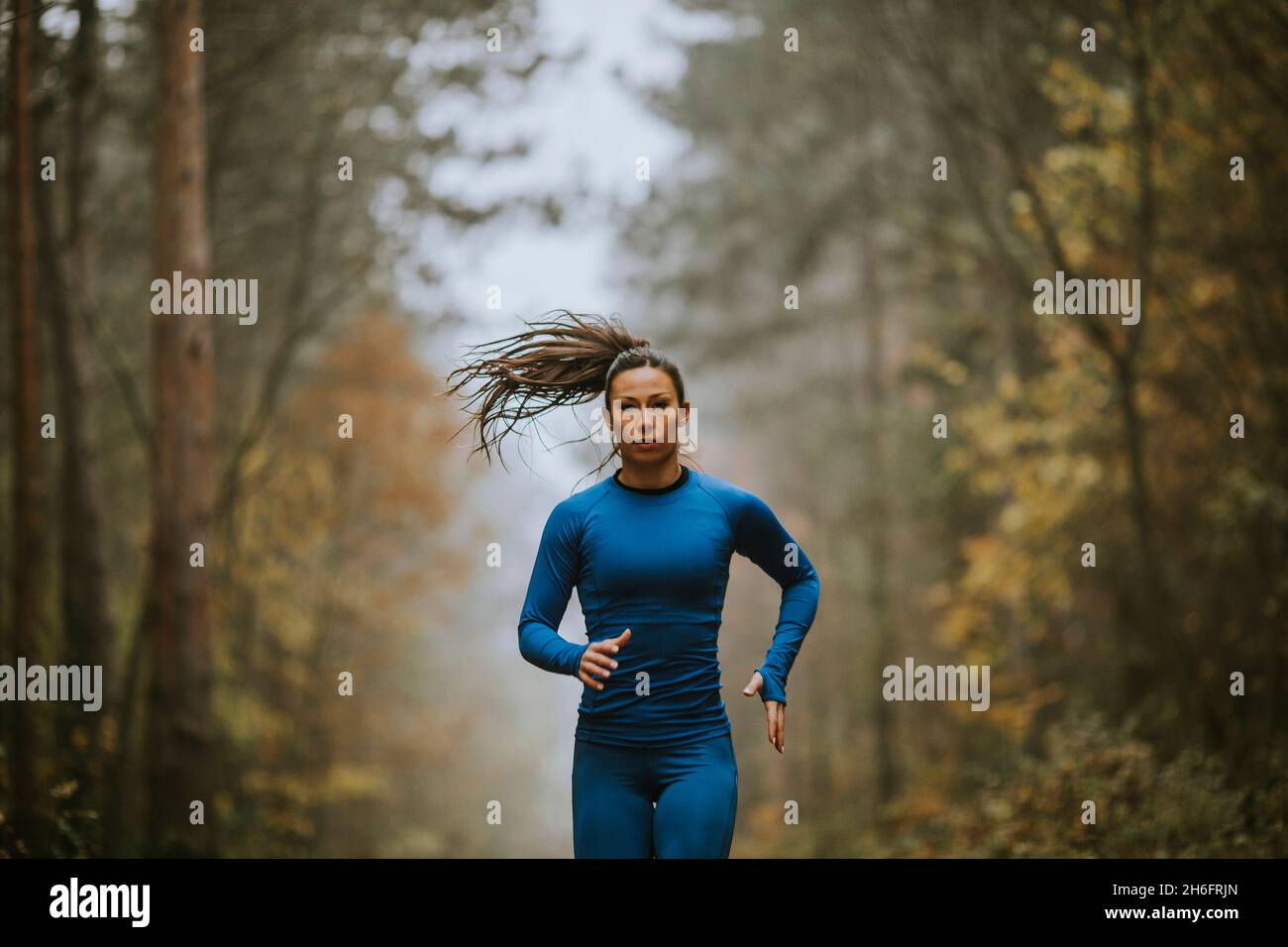 Giovane donna in tuta da pista blu che corre verso la macchina fotografica sul sentiero forestale in autunno Foto Stock