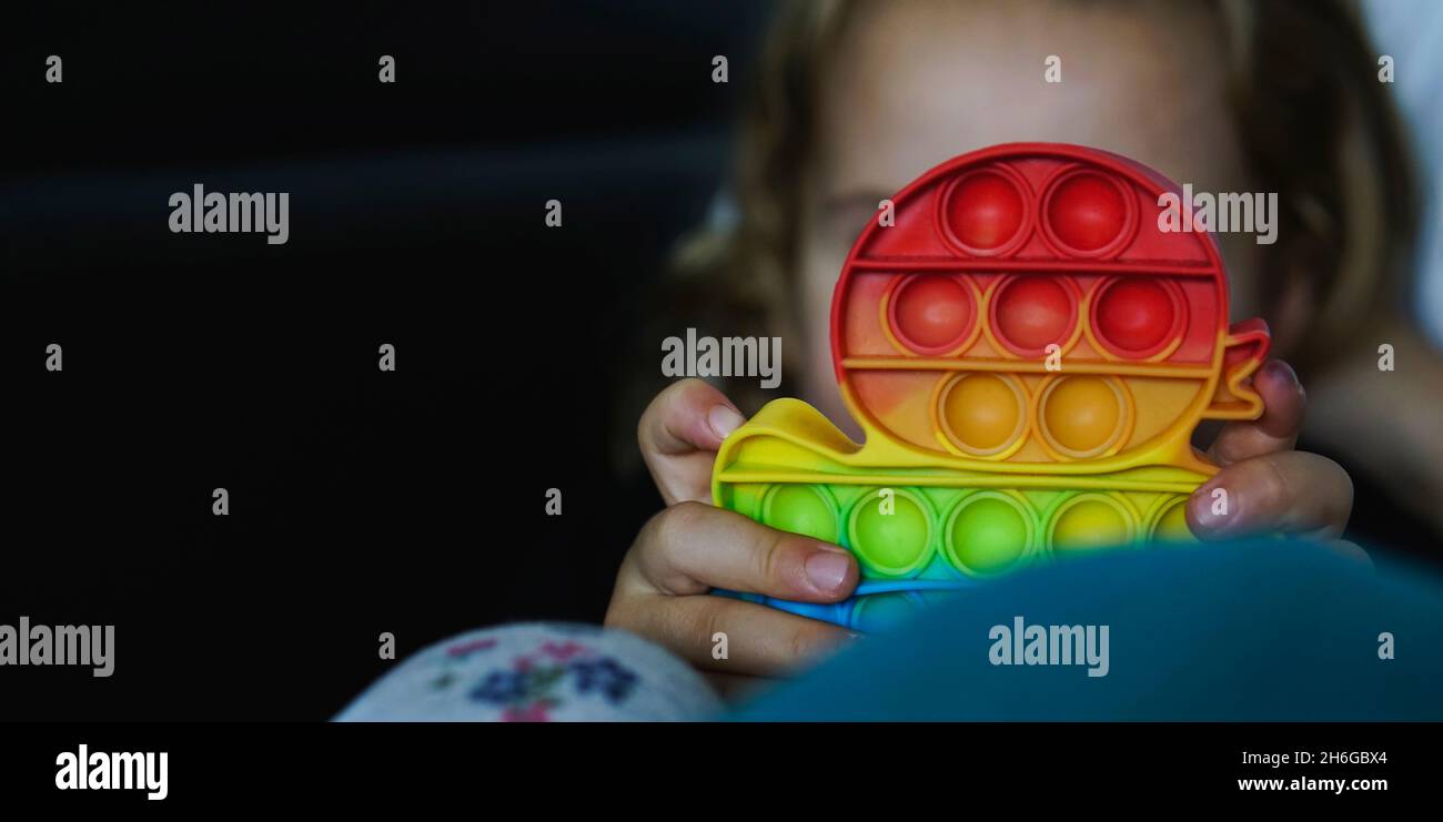 Piccolo bambino che gioca con il gadget pop-it arcobaleno. Popolare giocattolo antistress in silicone. Cultura giovanile e concetto di salute mentale. Giocattolo flessibile alla moda Foto Stock
