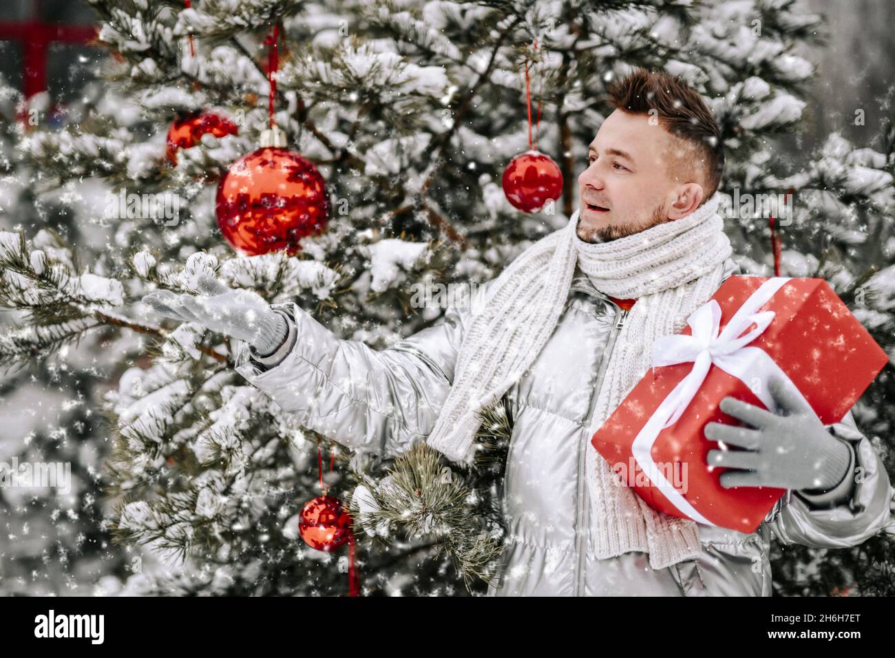 L'uomo felice bearded nel cappotto giù si alza all'albero di Natale decorato con giftbox rosso all'aperto che cattura i fiocchi di neve Foto Stock