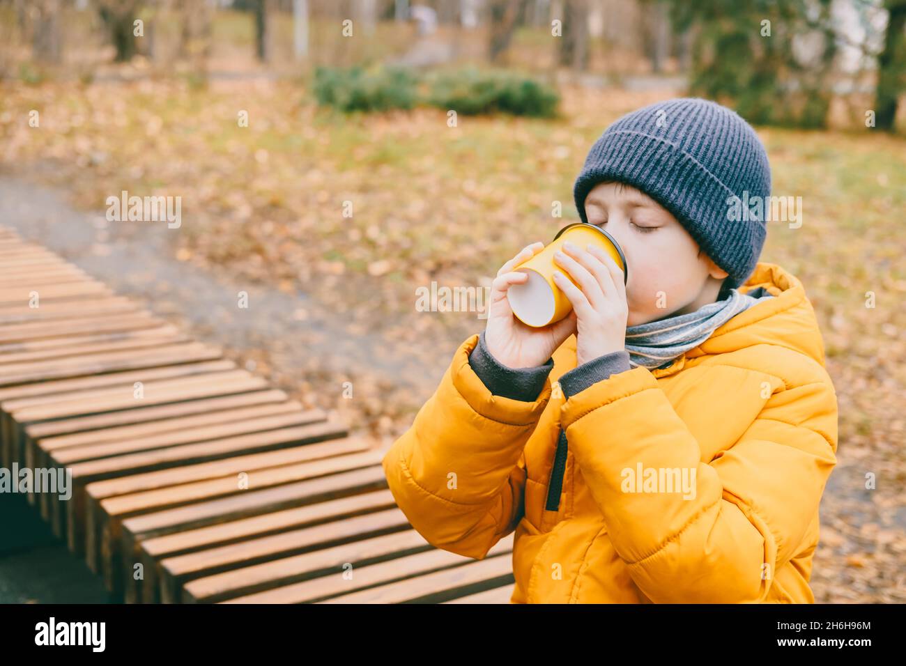 il ragazzo in una giacca gialla beve cioccolata calda da un bicchiere giallo, seduto su una panca, nel parco. il bambino si riscalda con un tè caldo durante una passeggiata Foto Stock