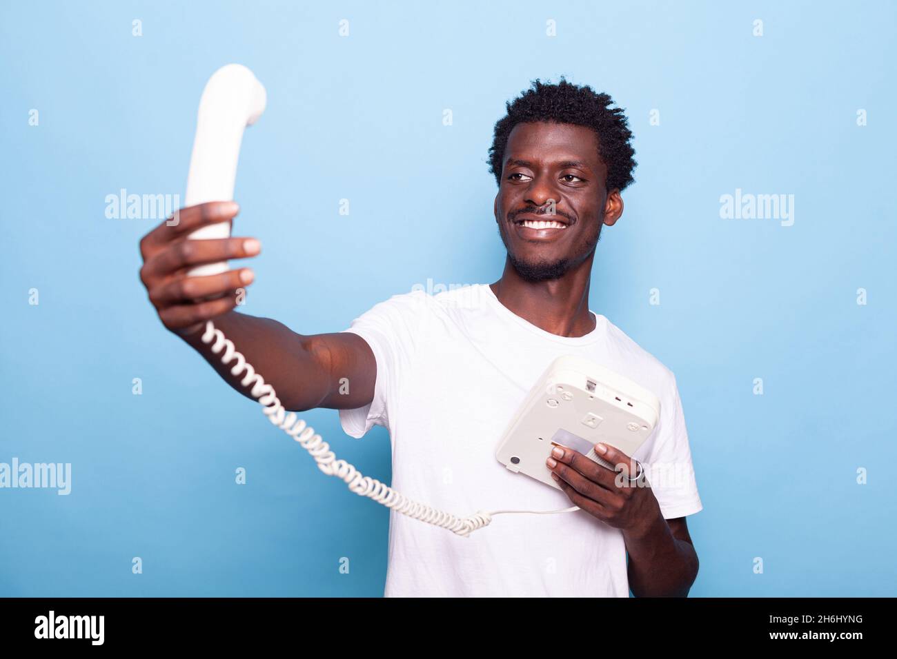Uomo afroamericano che finge di prendere selfie sul vecchio telefono fisso. Persona nera che si diverte con il telefono retrò, scherzando a scattare foto e sorridendo, usando il telefono vintage in mano Foto Stock