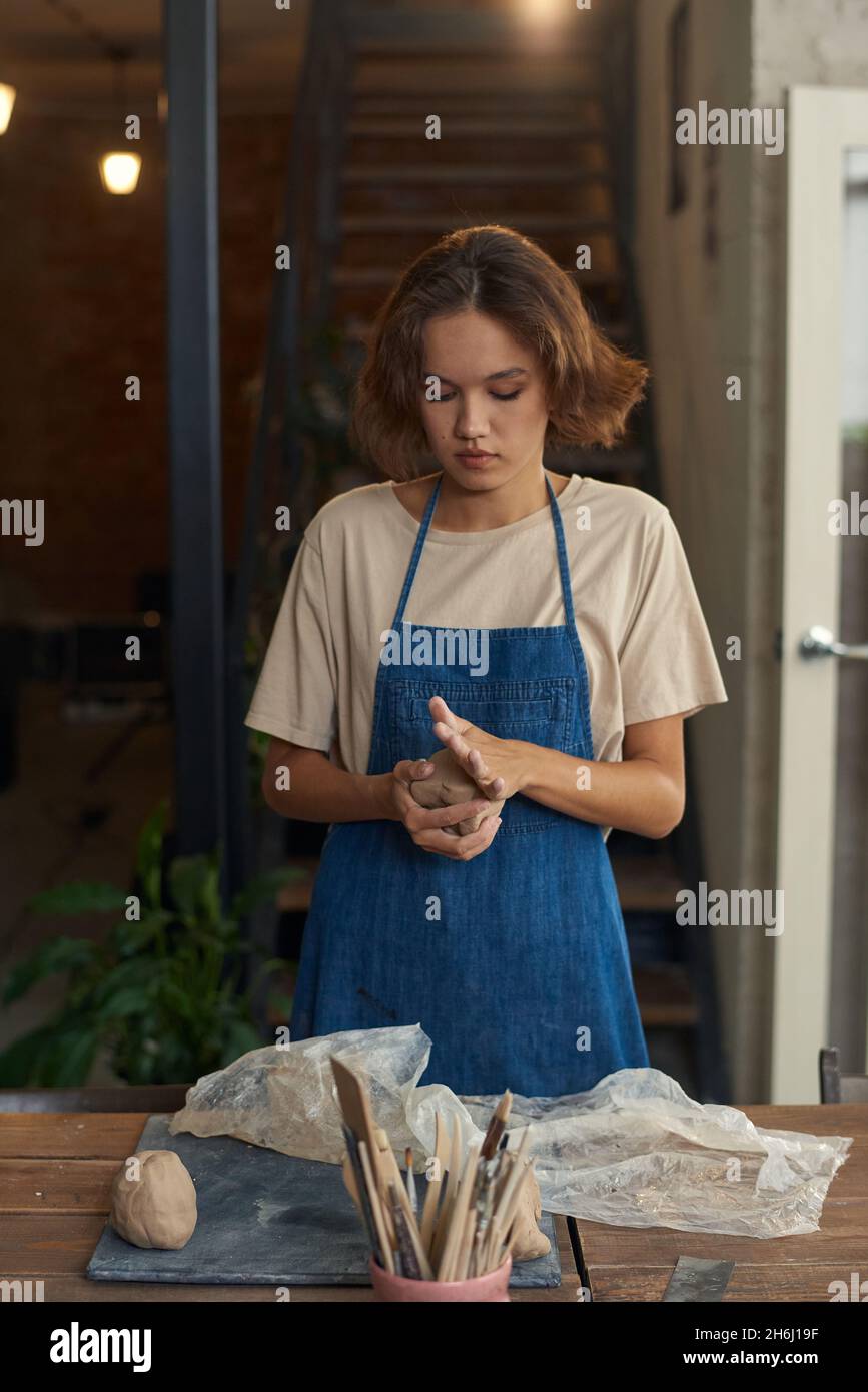 Giovane artigiano in abbigliamento da lavoro impastando il pezzo di argilla tra le mani mentre lo prepara per un'ulteriore lavorazione Foto Stock