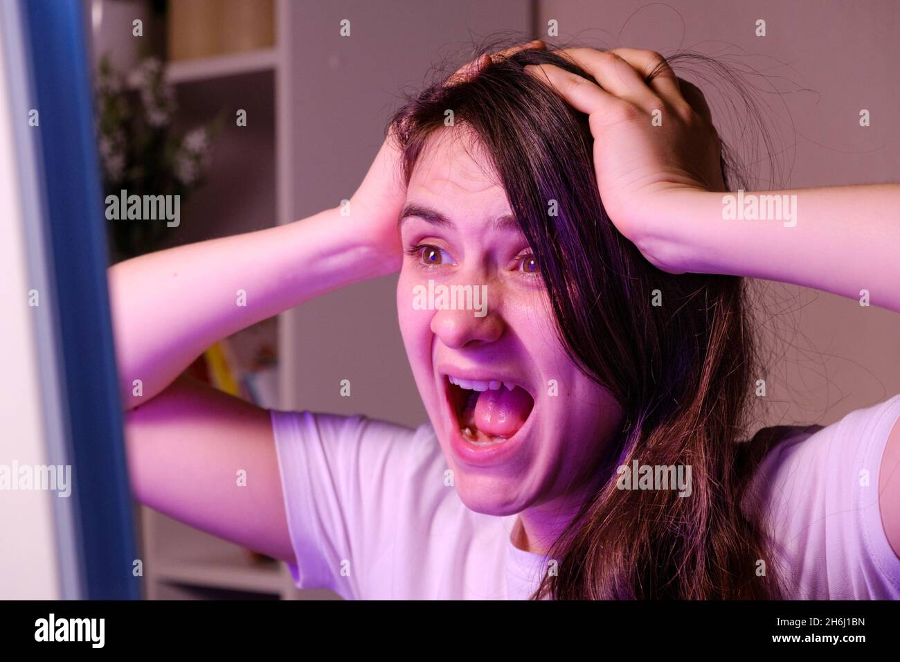 Una donna urla e afferra la testa con le mani mentre guarda una TV o uno schermo del computer. Foto Stock