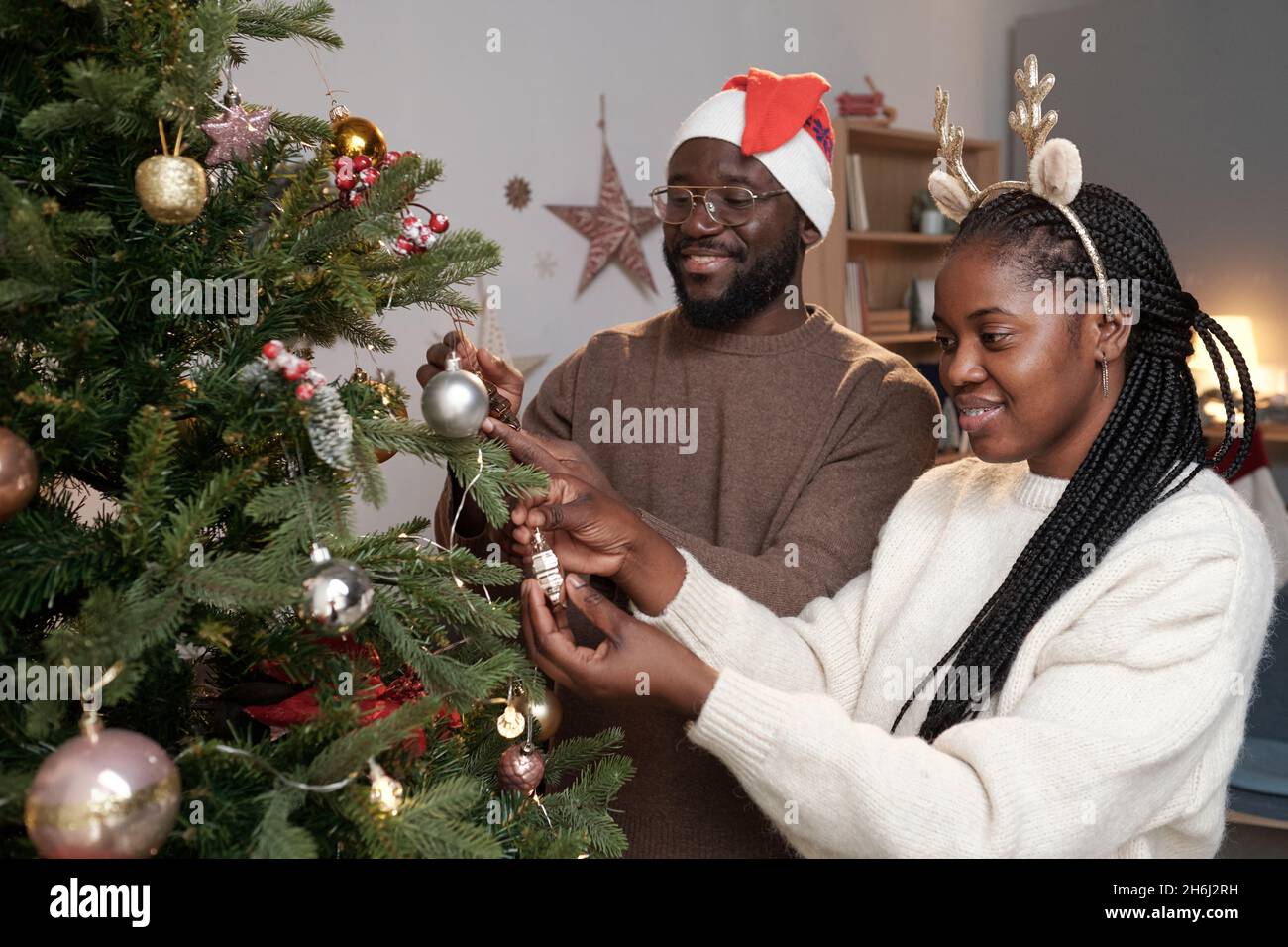 Giovane coppia africana in tradizionale cappello di Santa e fascia che decorano albero sempreverde con giocattoli in ambiente domestico Foto Stock