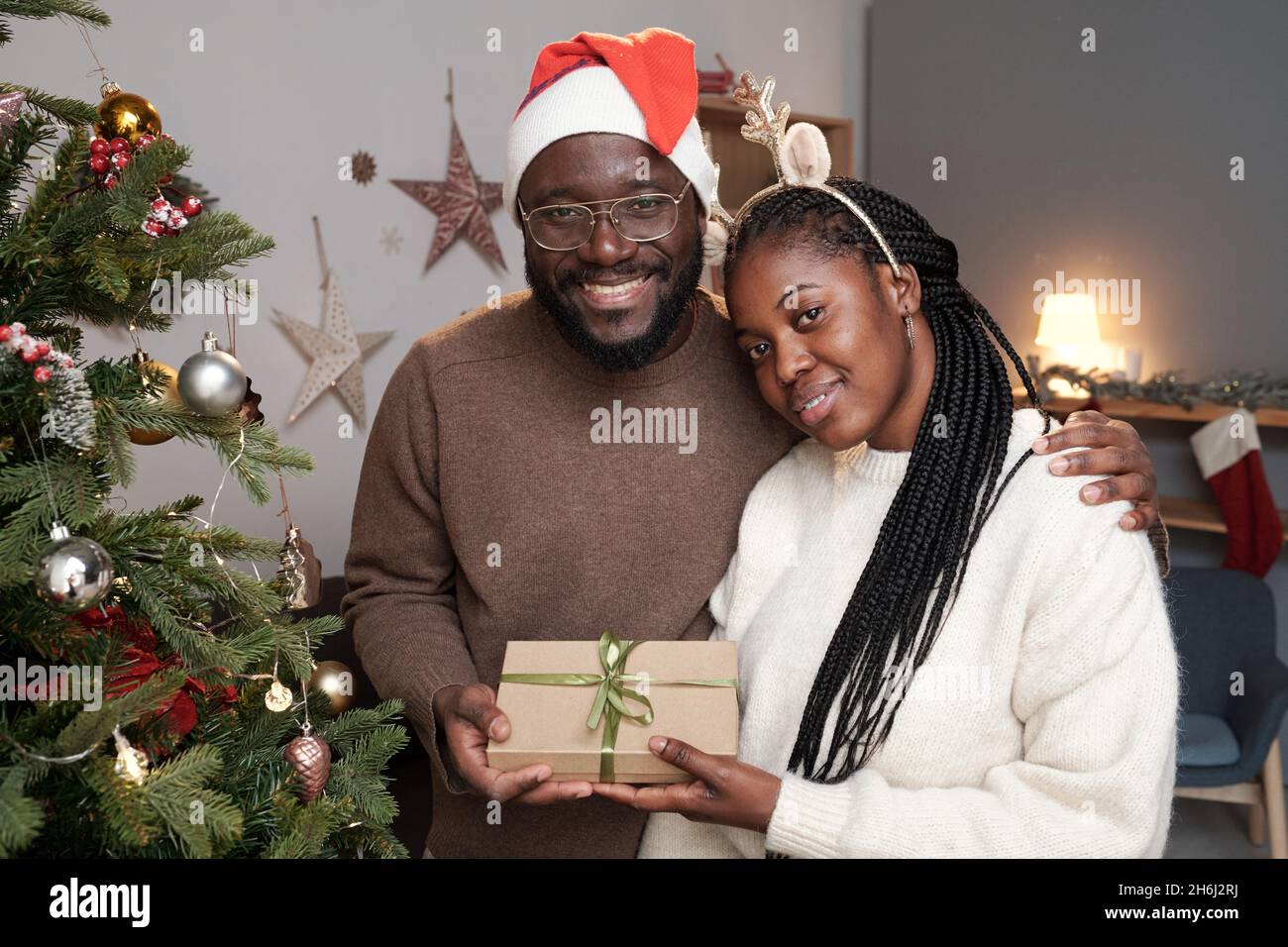 Felice giovane coppia africana in casualwear e cappellino e fascia di Natale in piedi di fronte alla macchina fotografica in soggiorno Foto Stock