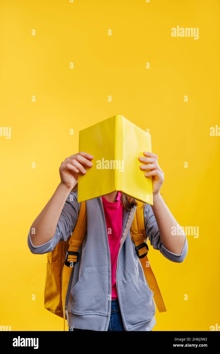 Ragazza adolescente felice che tiene zaino che copre la sua faccia con il libro. Ritratto studio su sfondo giallo con spazio vuoto per il testo. Foto Stock