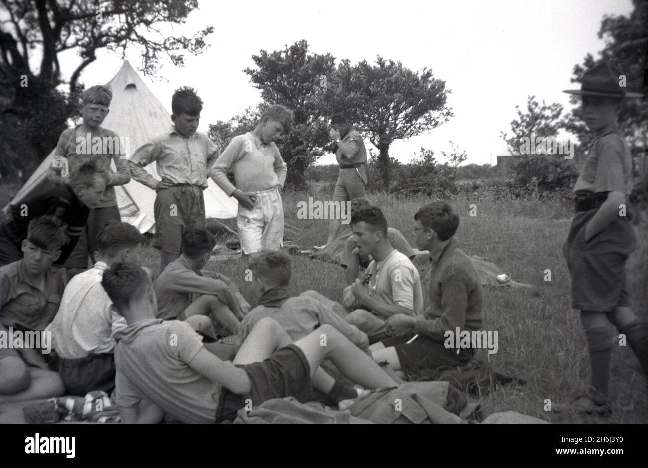 1938, storico, un campo scout, fuori in un campo, un gruppo di cub scout che ascolta un ragazzo più vecchio che gioca un ukulele, Felbridge, Surrey, Inghilterra, Regno Unito. Scouting iniziò nel 1908, dopo che il British Army Officer, Robert Baden-Powell tenne un campo sperimentale sull'isola di Brownsea, Poole Harbour, l'estate precedente. Circa 20 ragazzi hanno partecipato e sono stati insegnati una serie di abilità all'aperto, tra cui monitoraggio, vigili del fuoco, realizzazione di rifugi da rami, annodamento, cucina, salute e igiene, salvavita e pronto soccorso. Il famoso libro di Baden-Powell, 'Scouting for Boys', fu pubblicato per la prima volta nel 1908. Foto Stock