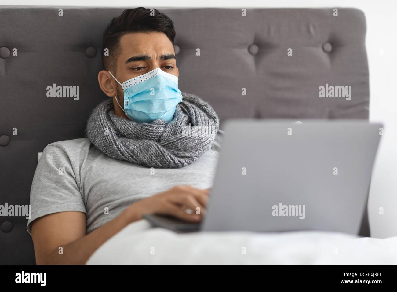 Malato giovane arabo uomo che indossa maschera medica lavoro con laptop in letto Foto Stock