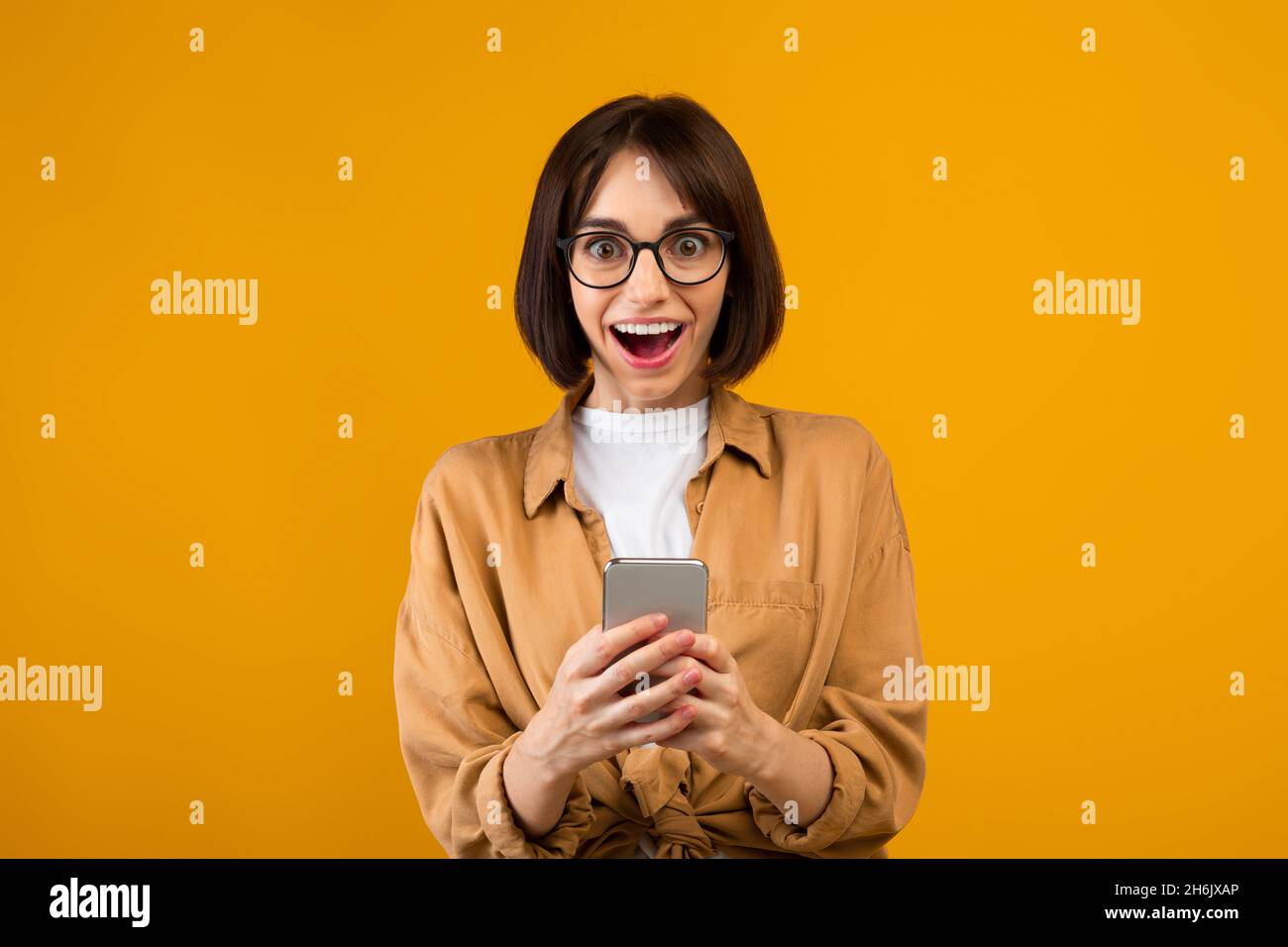 OMG, incredibile. Sorprende la donna che tiene e utilizza lo smartphone, ricevendo messaggi, sfondo giallo studio Foto Stock