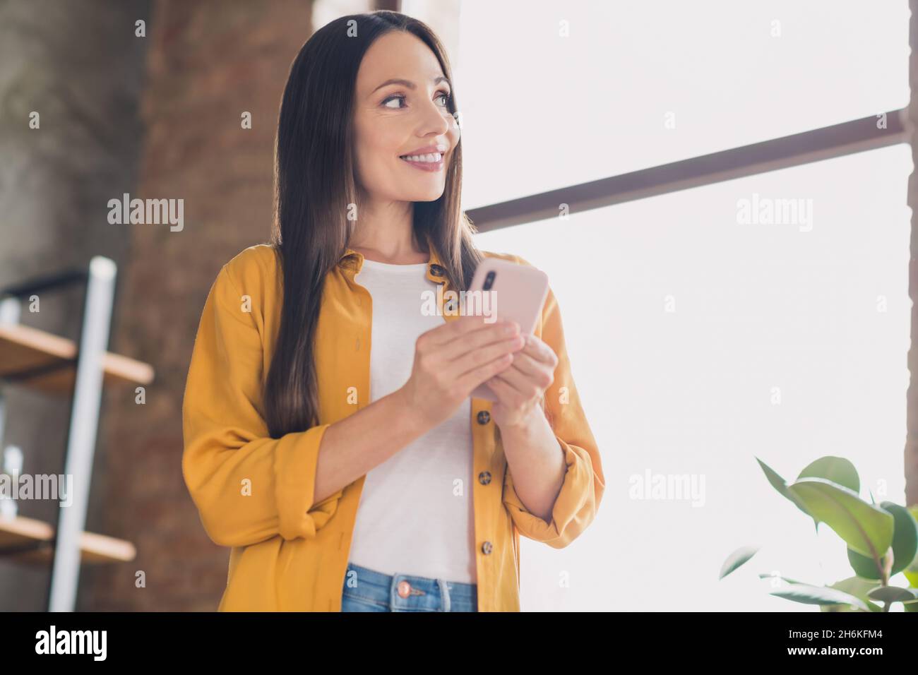 Foto di affascinante signora giovane sognante vestita camicia gialla digitando dispositivo moderno guardando finestra sorridente casa stanza interna Foto Stock