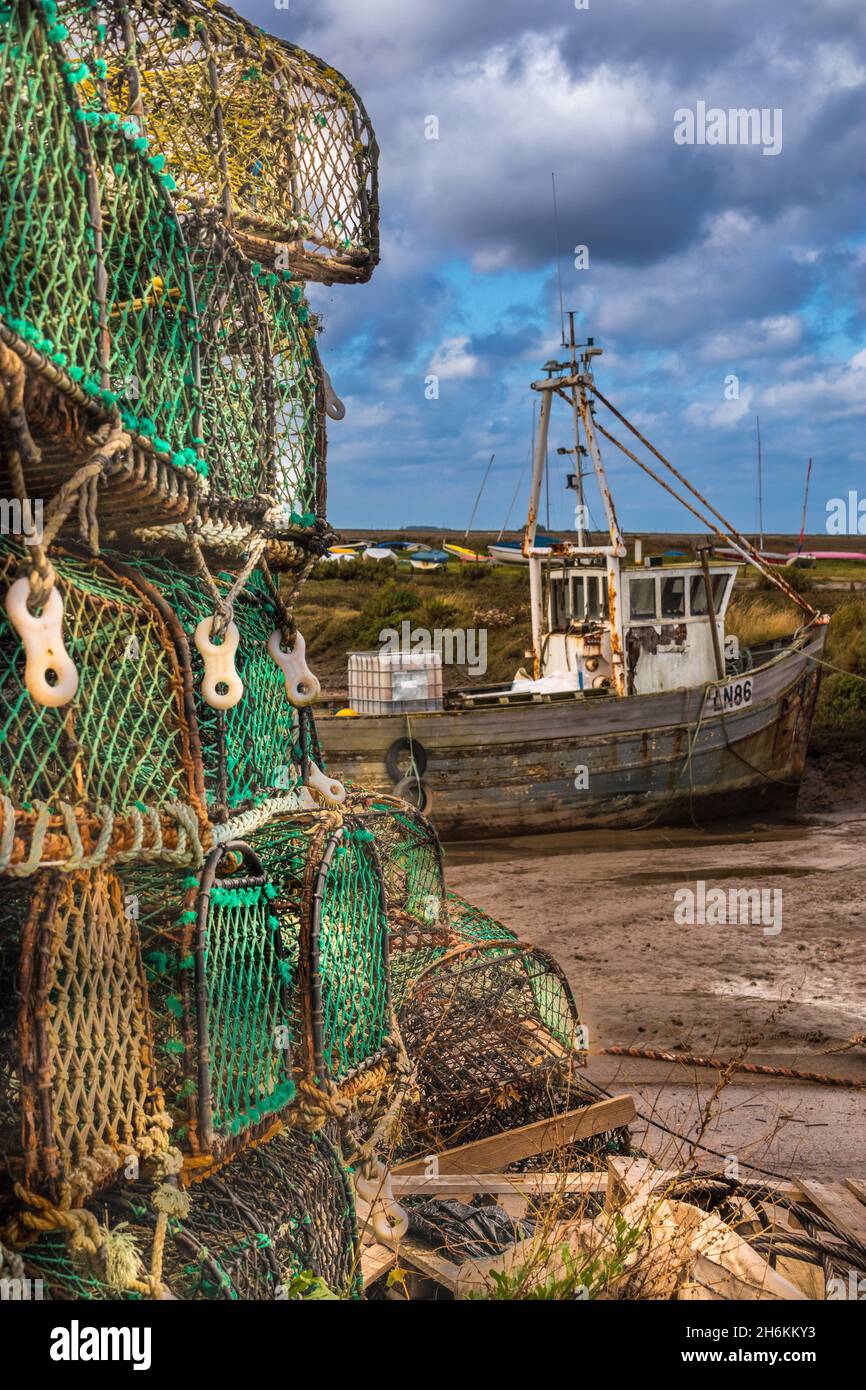 Stack di gabbie di granchio in primo piano con vecchia barca da pesca sul fango a bassa marea a Brancaster Staithe Harbour Nord Norfolk Inghilterra Foto Stock