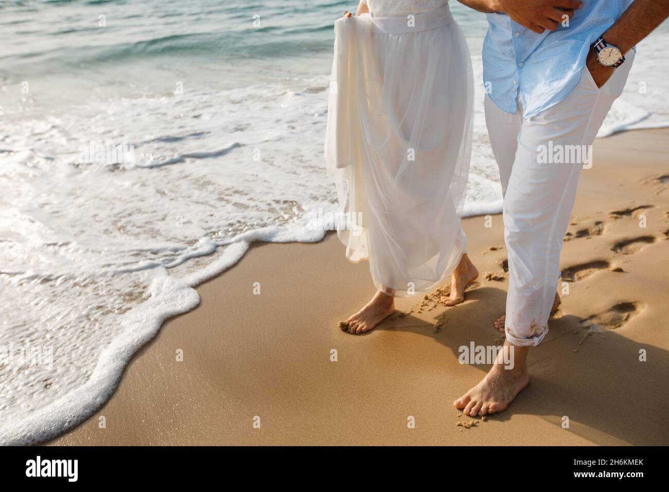 Luna di miele degli sposi novelli. Sposa a piedi nudi in abito bianco e sposi in pantaloni bianchi camminando sulla romantica spiaggia dell'oceano. La giovane coppia si innaffia sulla sabbia dorata. Foto Stock