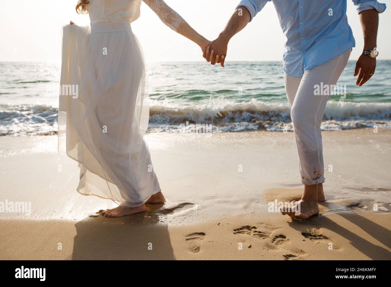 Luna di miele degli sposi novelli. Sposa a piedi nudi in abito bianco e sposi in pantaloni bianchi camminando sulla romantica spiaggia dell'oceano. La giovane coppia si innaffia sulla sabbia dorata. Foto Stock