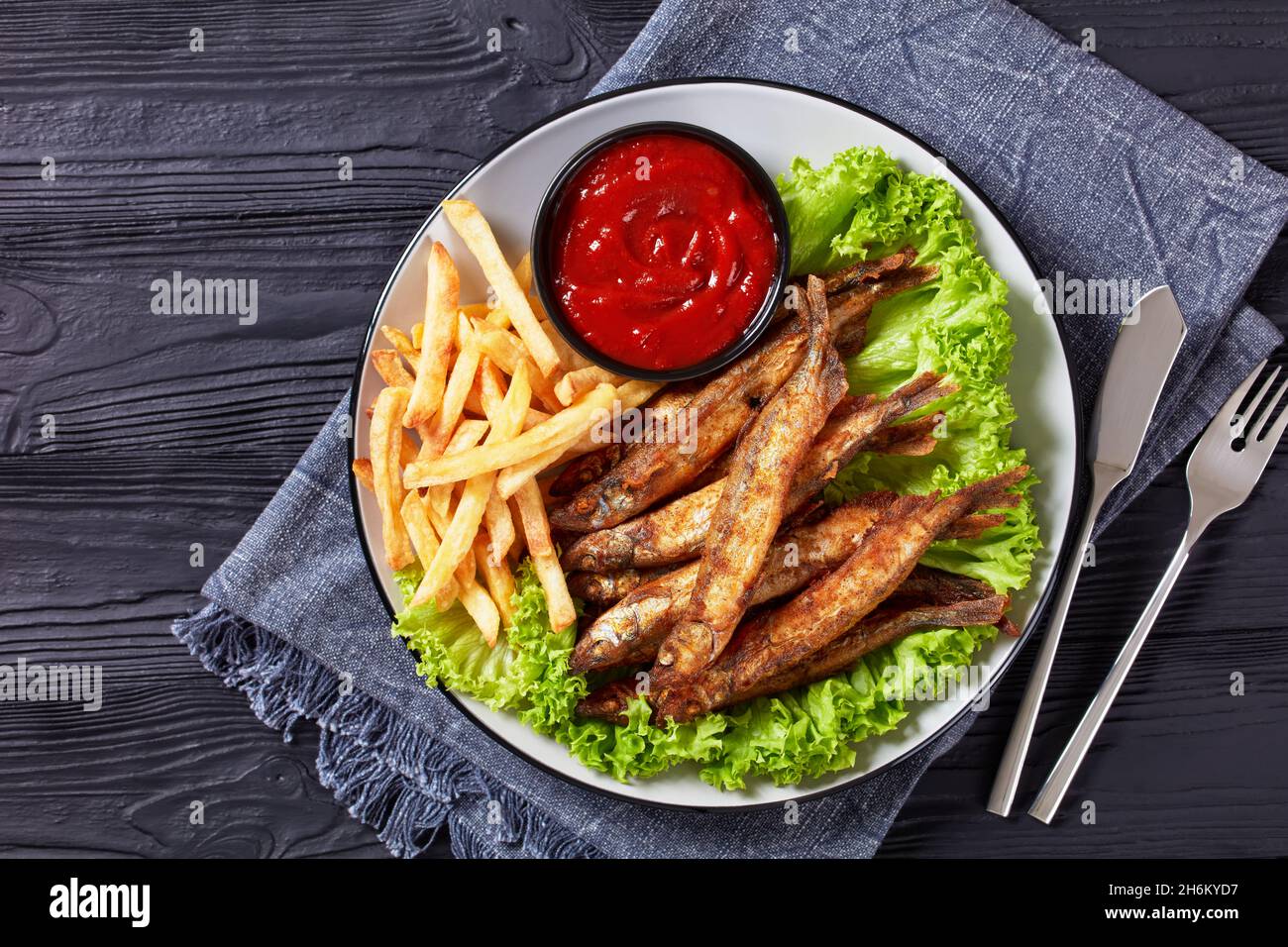 pesce fritto croccante di capelin con lattuga fresca, patatine fritte e salsa di pomodoro su un piatto su un tavolo di legno nero testurizzato, piatto, spazio libero Foto Stock