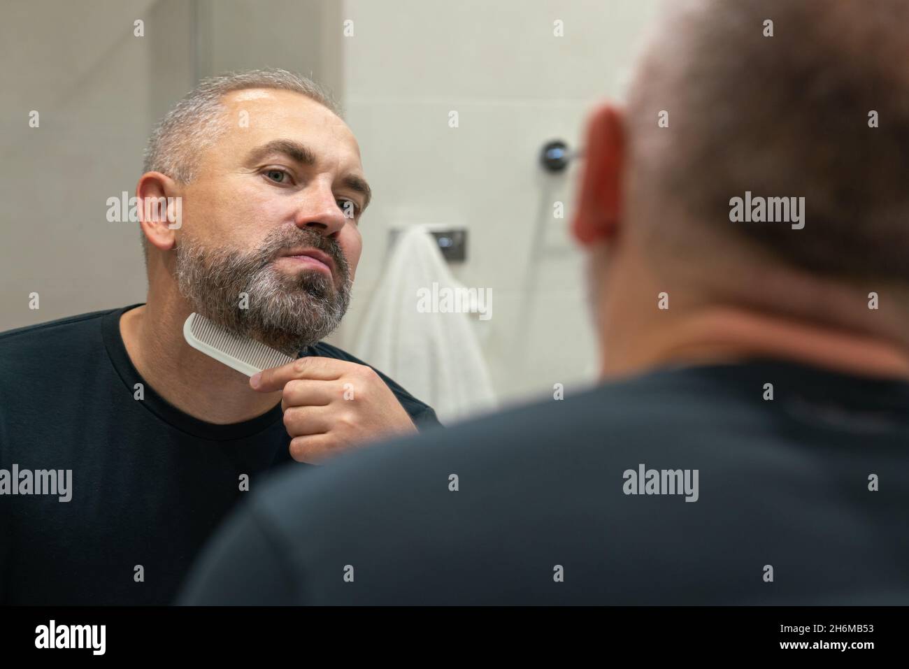 Un bell'uomo di mezza età che gli spazzolava la barba la mattina in bagno guardando nello specchio Foto Stock