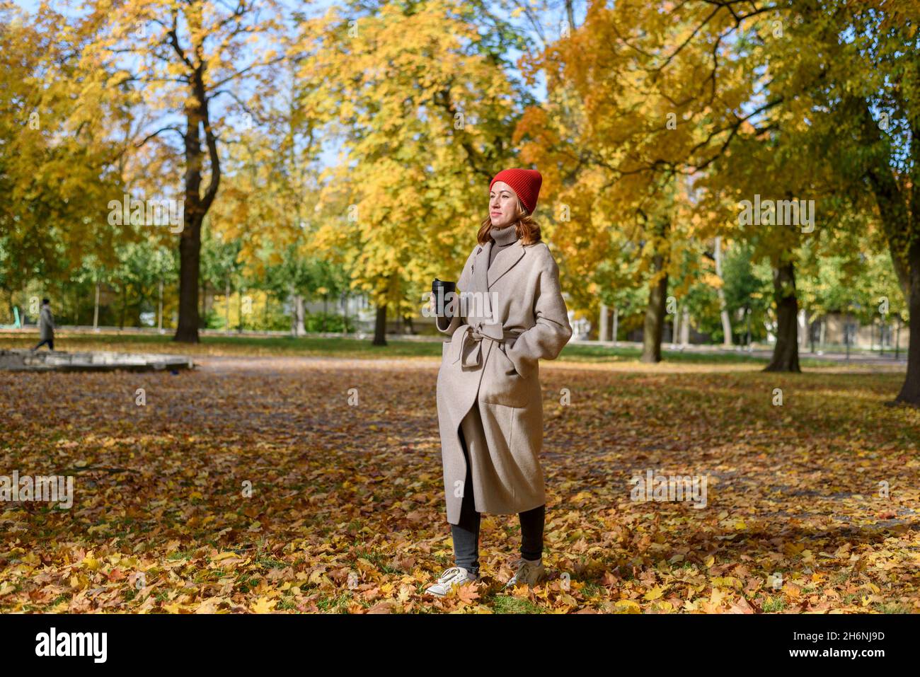 Donna in cappotto di lana e cappello di maglia rossa a piedi e bere caffè da tazza riutilizzabile al parco autunnale Foto Stock