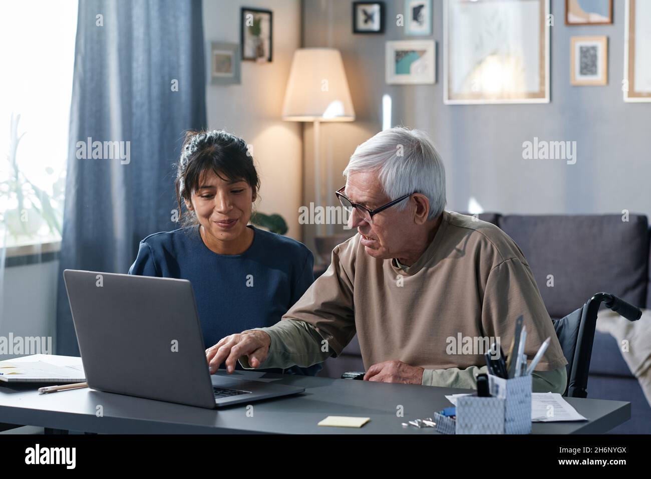 Uomo anziano imparando ad usare il computer con l'aiuto della donna mentre si siedono al tavolo insieme nella stanza Foto Stock