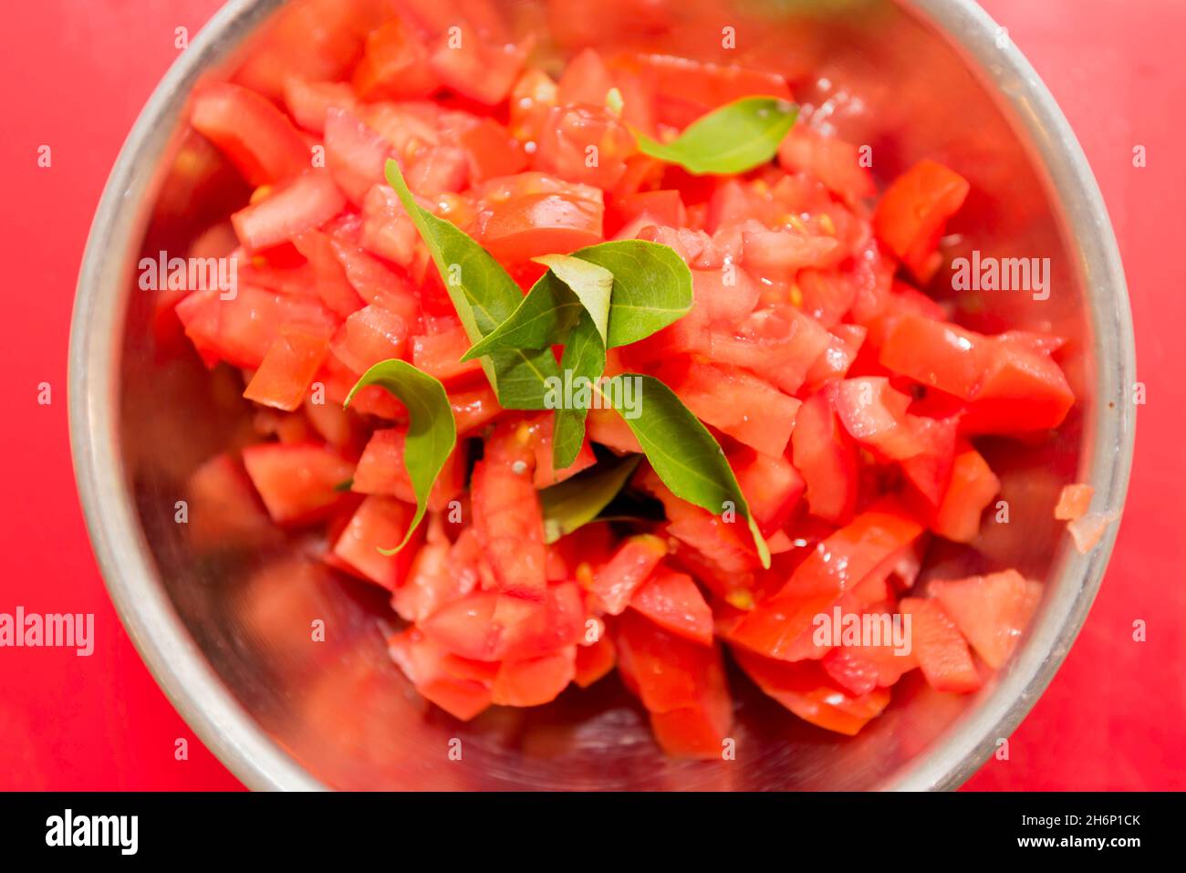 Guardando giù su una ciotola di acciaio inossidabile riempita con pomodori rossi tritati brillanti e alcune foglie di curry, su un bordo rosso di tritare Foto Stock
