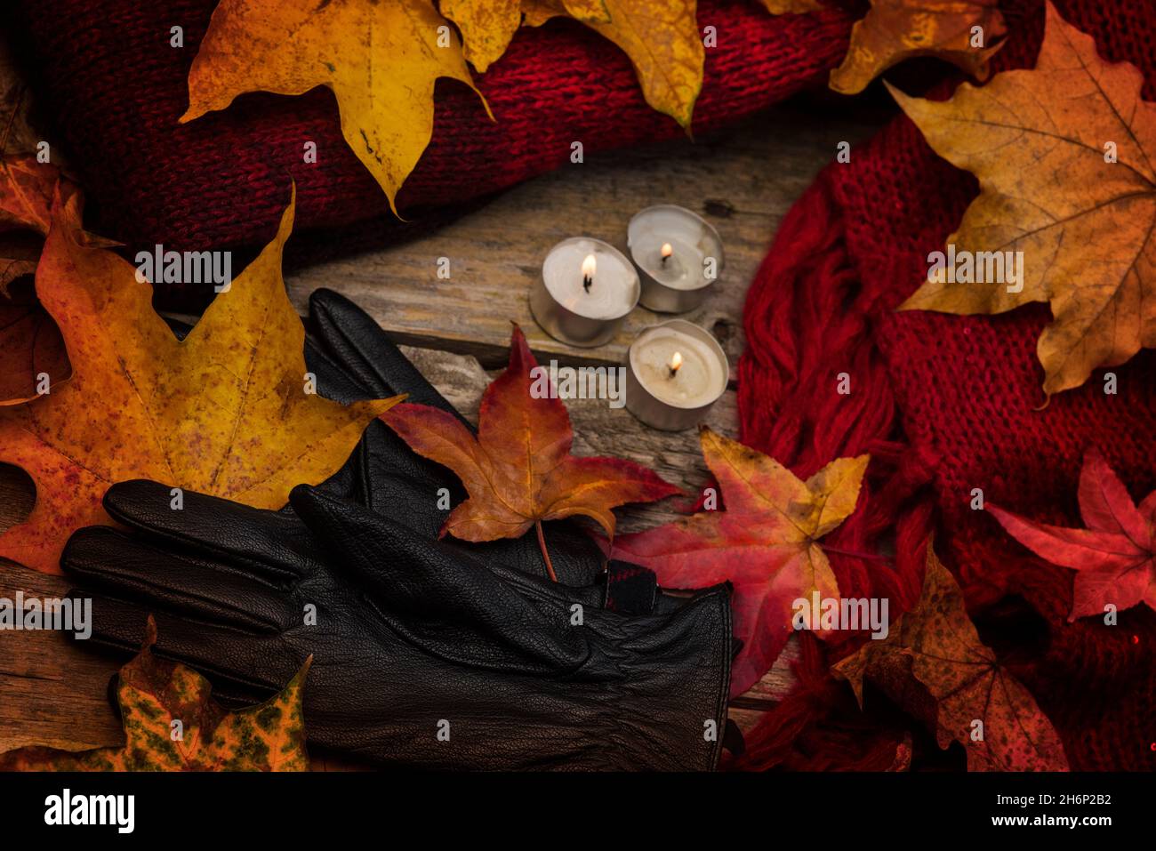 Moda autunno/autunno. Jumper in lana spessa, sciarpa e guanti in pelle sintetica con foglie cadute sparse e tealights. Stagionale. Clima freddo/freddo. Foto Stock