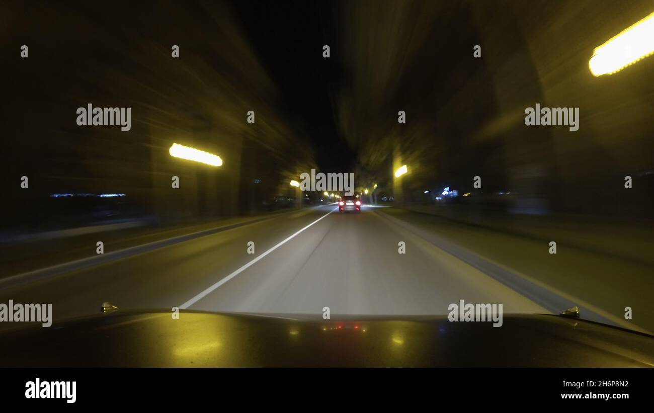 I movimenti dell'auto sulla strada stretta della città di notte. Un'auto sta guidando avanti. Immagine sfocata di alberi e luci sui bordi della strada Foto Stock