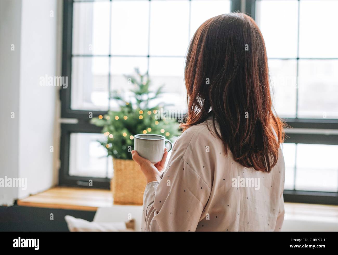 Giovane adulta quarant'anni donna con capelli scuri nel pigiama accogliente con tazza di tè in soggiorno con albero di Natale, gente da dietro Foto Stock