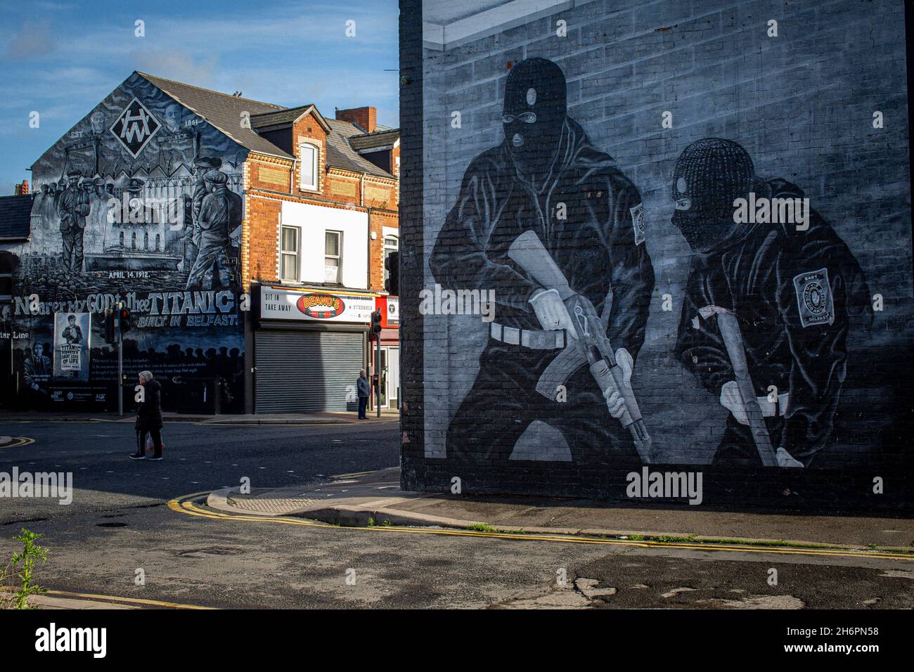 Persone che camminano davanti a un murale paramilitare lealista con uomini mascherati che tengono mitragliatrici sulla Newtownards Road a Belfast, Irlanda del Nord. Foto Stock