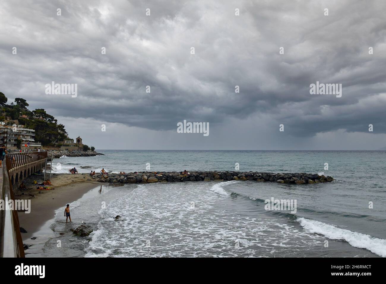 Vista panoramica sulla riva del mare con frangiflutti rocciosi e la Cappella ('Cappelletta') sullo sfondo durante una tempesta estiva, Alassio, Savona, Liguria Foto Stock