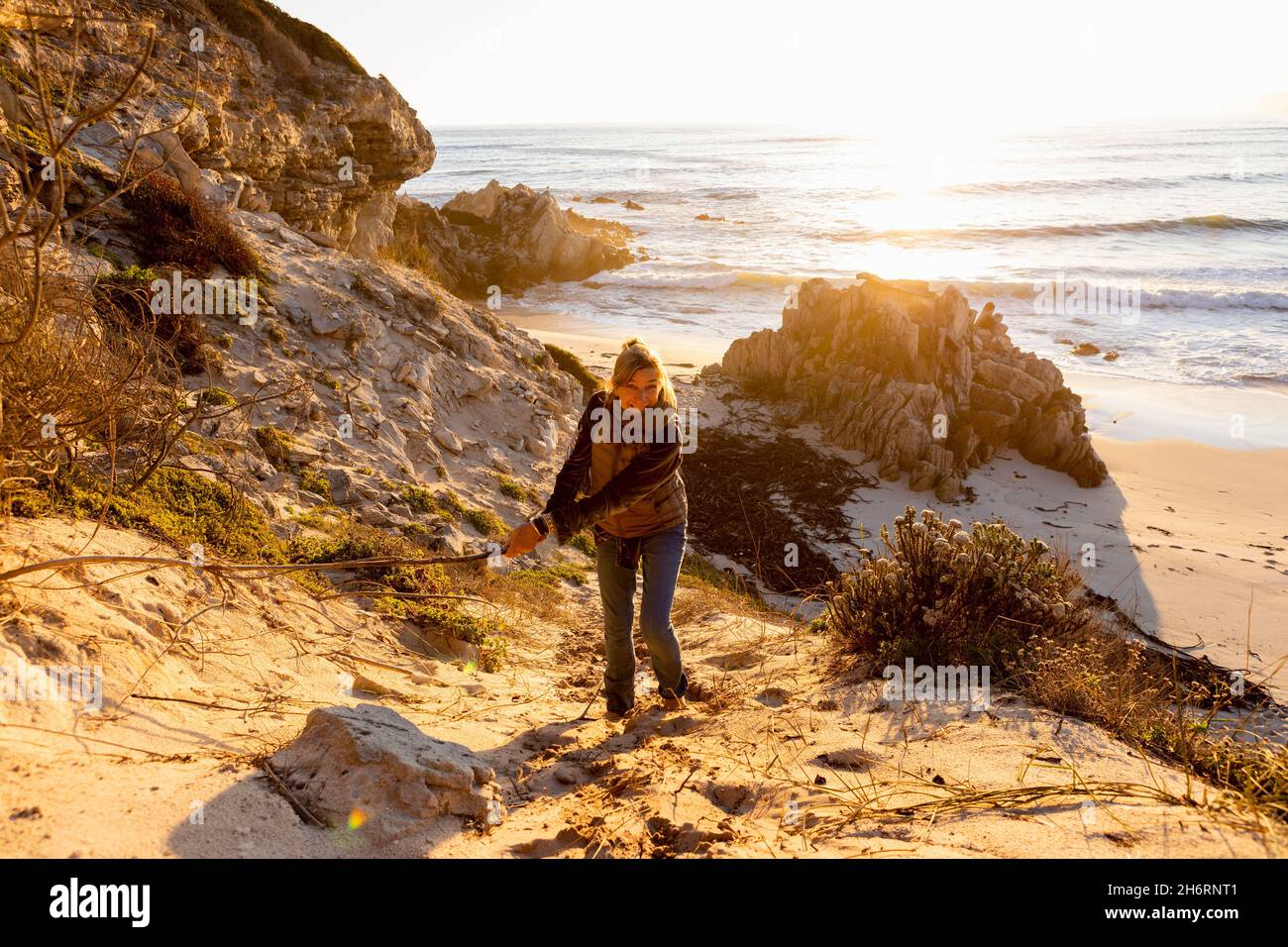 Donna in piedi sulle scogliere sopra una spiaggia ondeggiante un lungo bastone e ridente Foto Stock