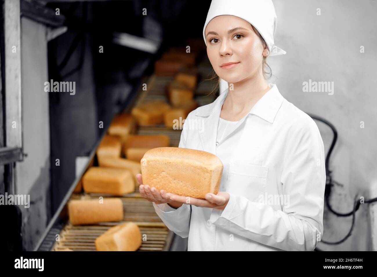 Ritratto di donna panettiere che tiene il pane fresco in mani sullo sfondo trasportatore automatico di panetteria alimentare fabbrica. Foto Stock