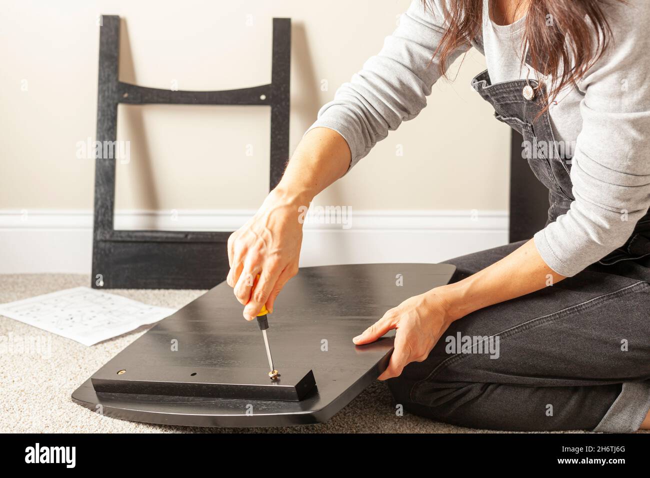 Concetto di assemblaggio di mobili fai da te che include una persona caucasica su pavimento di moquette con pezzi di mobili, cercando di montarlo. Tiene una vite gialla Foto Stock