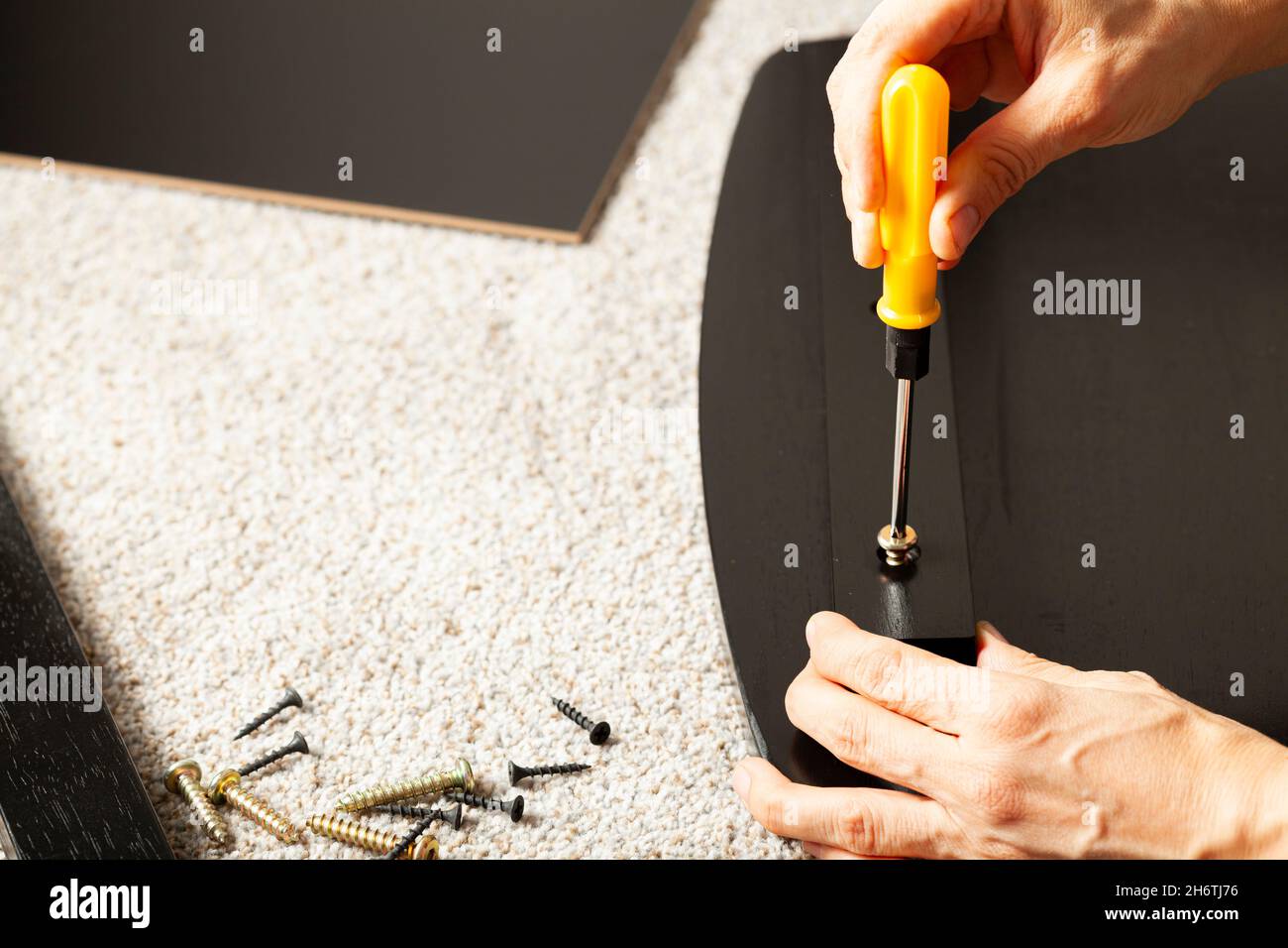 Concetto di assemblaggio di mobili fai da te che include una persona caucasica su pavimento di moquette con pezzi di mobili, cercando di montarlo. Tiene una vite gialla Foto Stock