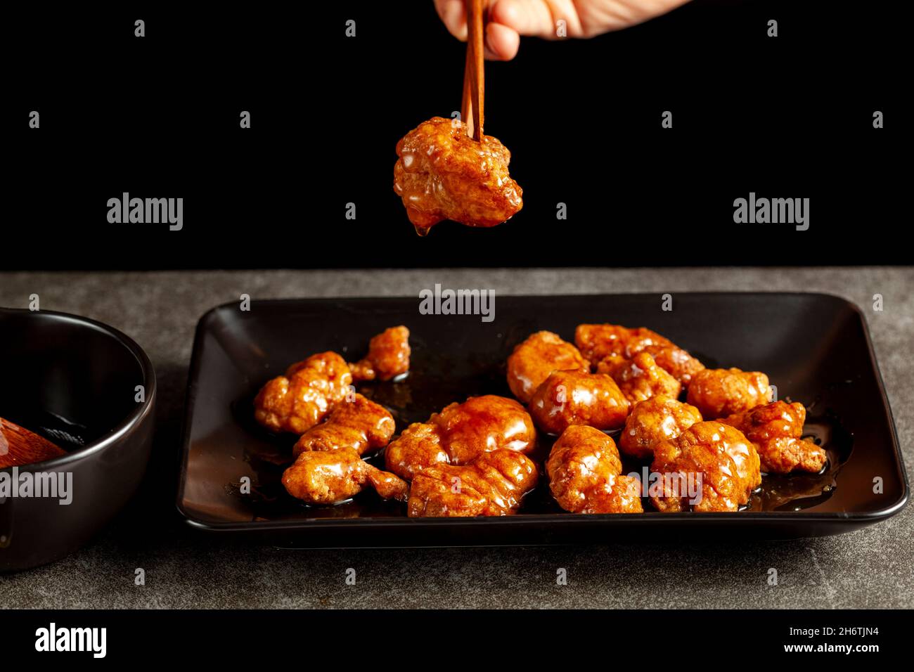 Una persona sta mangiando il pollo d'arancia del mandarino delizioso, un piatto americano cinese usando i bastoncini di legno. La salsa viene servita in una ciotola di porcellana. bac. Scuro Foto Stock