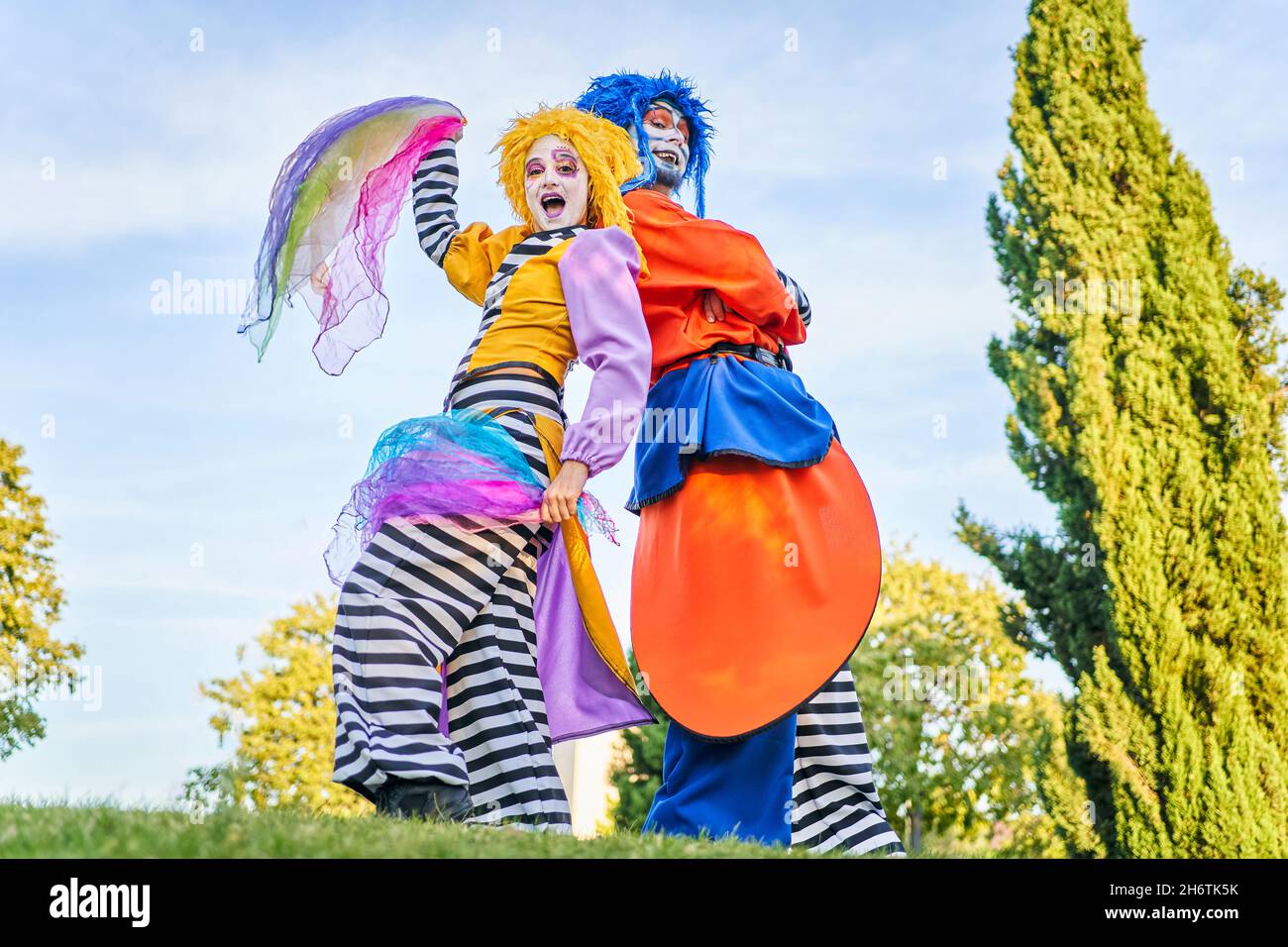 Vista laterale di allegro harlequins maschio e femmina in parrucche e costumi colorati in piedi sul prato con ritorno a casa durante il festival nel parco verde, in giorno di sole Foto Stock