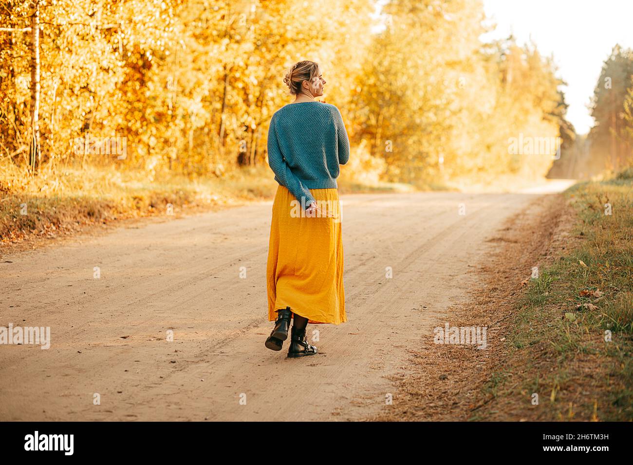 Foto di giovane donna passeggiate lungo la strada tra foresta autunno indossare gonna lunga gialla e maglione blu, vista posteriore, godere di solitudine, tranquillità, silenzio. Foto Stock