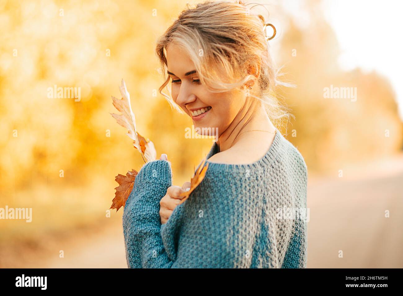 giovane bella donna, bionda con i capelli tirati indietro, indossando il maglione blu, sorridendo sullo sfondo di fogliame giallo, tenendo le foglie d'autunno dentro Foto Stock