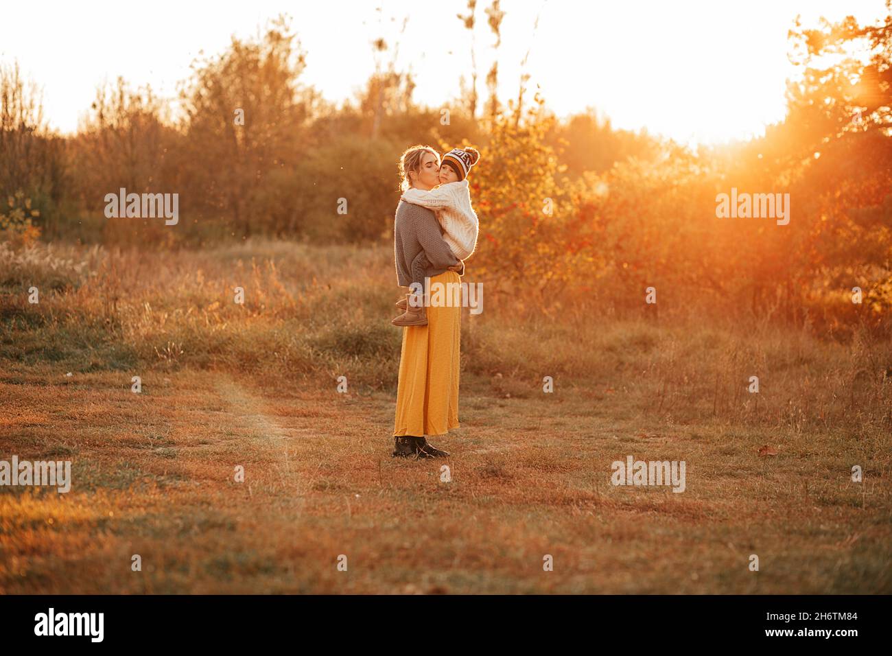 Una foto a tutta lunghezza di una giovane donna che cammina attraverso il campo d'autunno con il bambino in braccio. Raggi di sole tramontare, tramonto, tristezza. Stagione di caduta. Singola Foto Stock