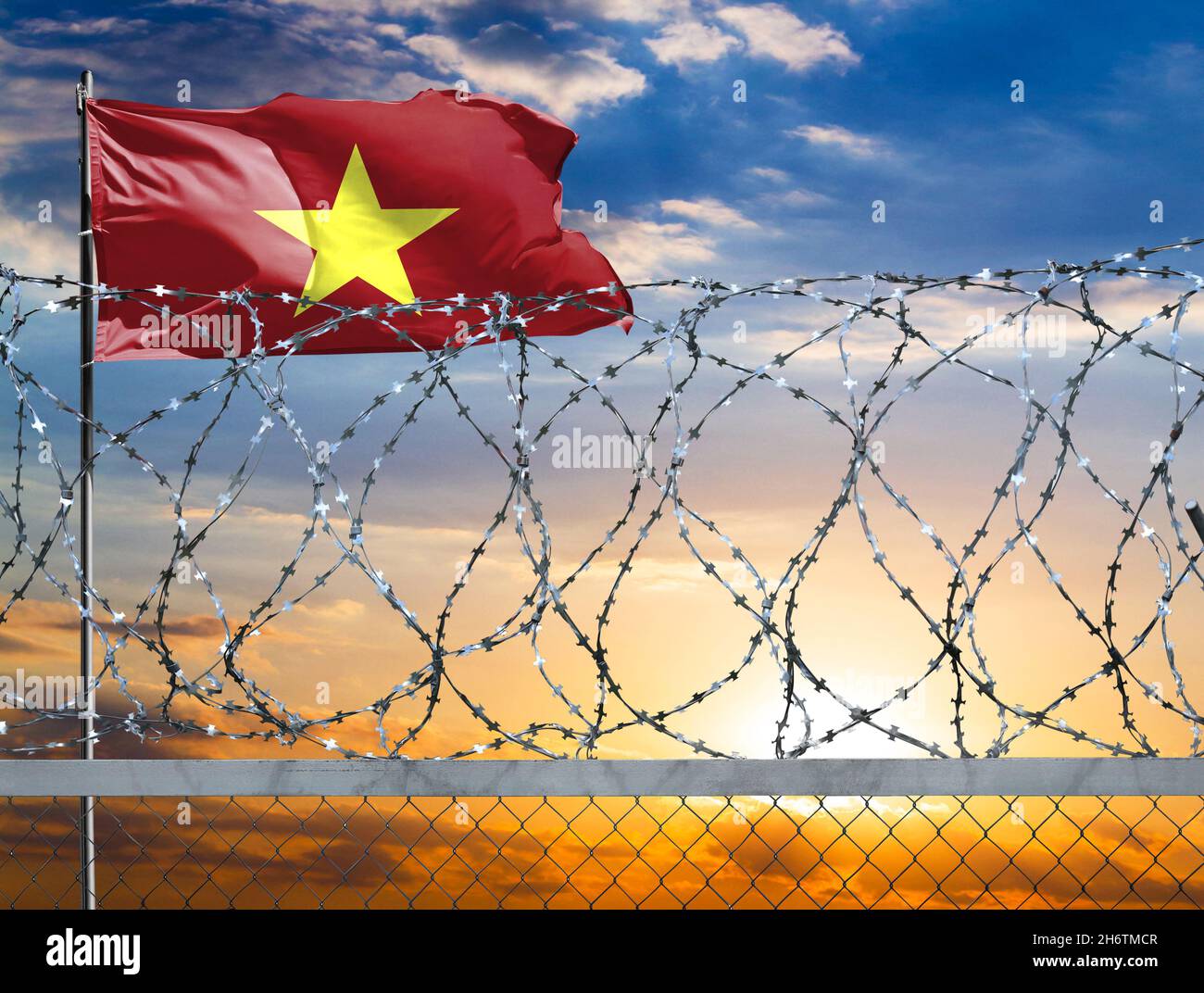 Una recinzione con filo spinato sullo sfondo di un cielo colorato e un flagpole con la bandiera del Vietnam protegge il confine dello stato da mig illegale Foto Stock