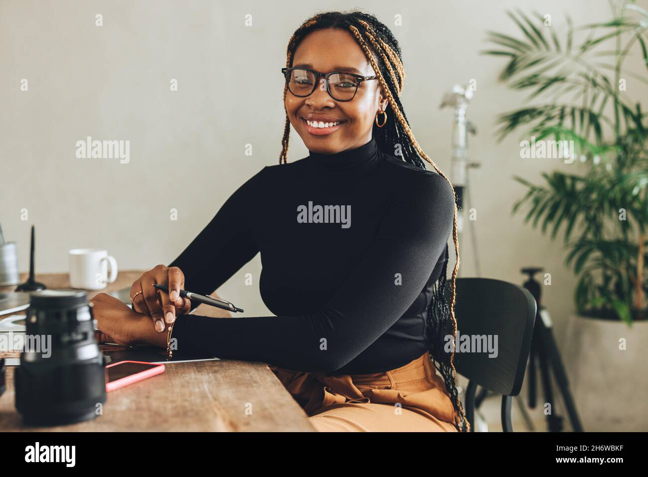 Giovane donna creativa sorridente alla macchina fotografica nel suo ufficio domestico. Felice fotografo femmina che tiene una penna stilo mentre si siede alla sua scrivania. Giovane freelan Foto Stock