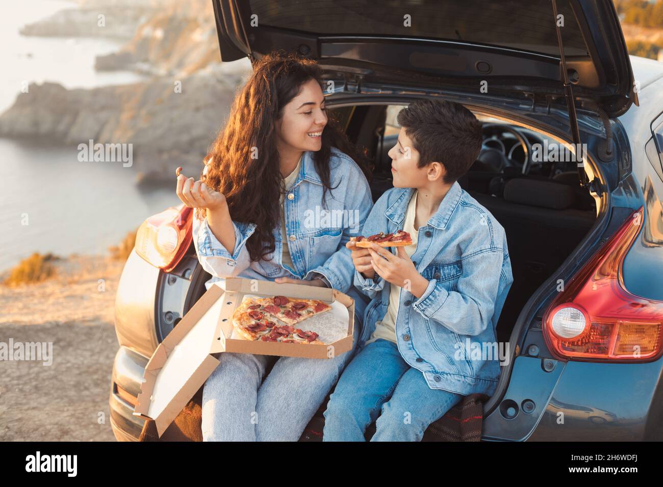 Una giovane madre e suo figlio adolescente in abiti denim seduti nel tronco aperto di una macchina e mangiare pizza Foto Stock