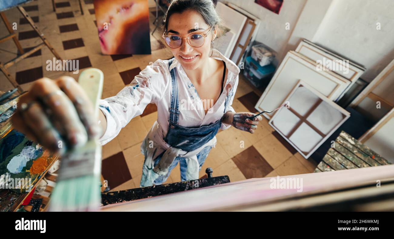 Allegro artista pittura su una tela nel suo studio. Vista ad angolo alto di un giovane pittore fantasioso sorridente mentre lavora. Artista creatrice maki femminile Foto Stock