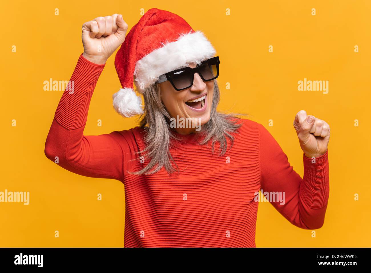 Felice donna di mezza età grigio-capelli in rosso santa cappello e occhiali da sole che ballano, fa movimenti alla musica, sorride positivamente, essendo in alto spirito. Spensierate danze mature della signora isolate su giallo Foto Stock