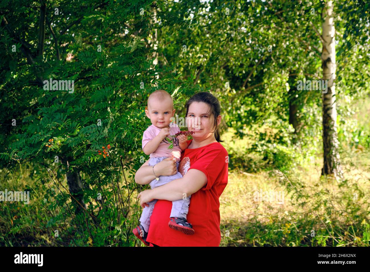 vista di mezza lunghezza di una giovane donna con un bambino nella foresta in piedi sullo sfondo di alberi verdi betula pendula e sorbus aucuparia Foto Stock