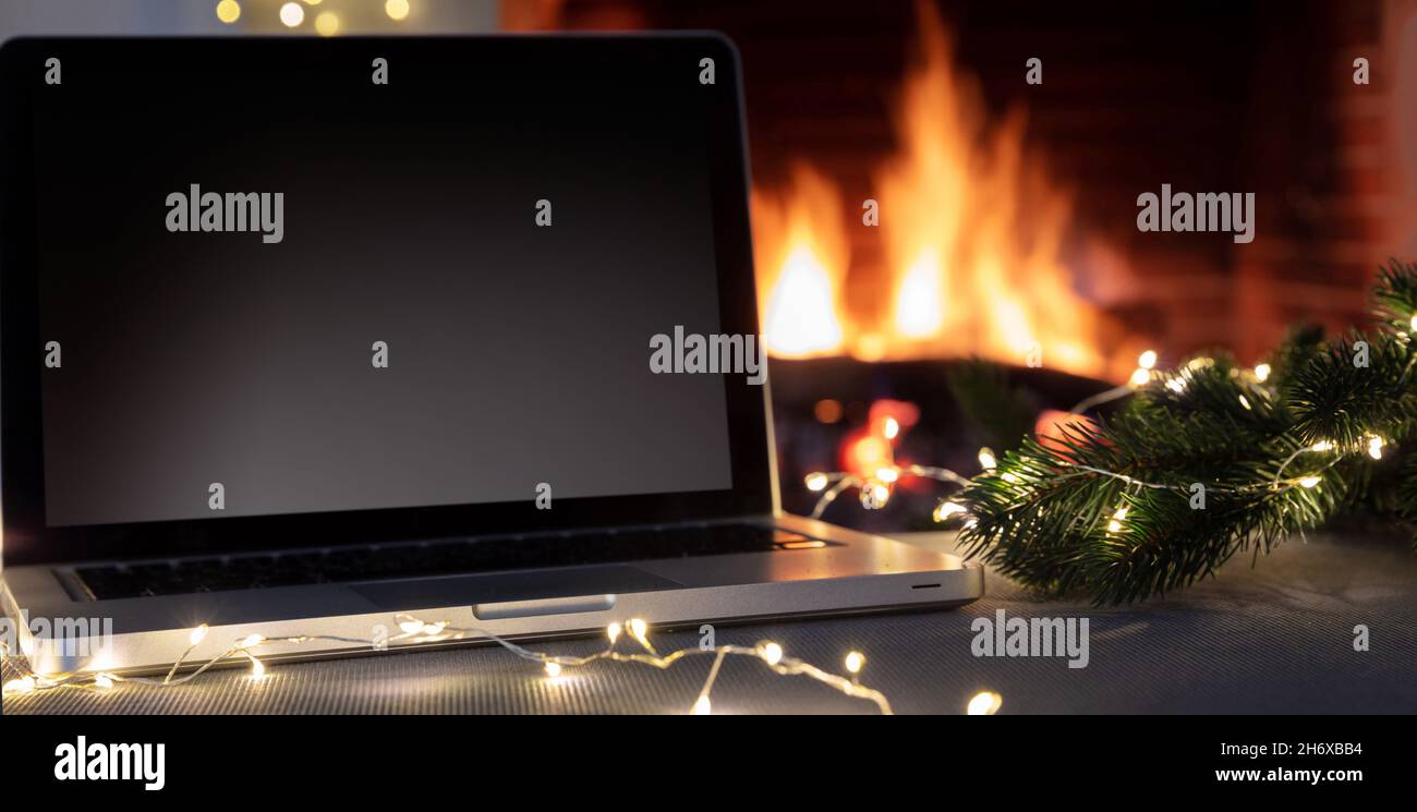 Concetto di ufficio domestico di Natale. Computer portatile con schermo vuoto su sfondo camino acceso, schermo nero vuoto del computer. Vacanze invernali accogliente casa Foto Stock